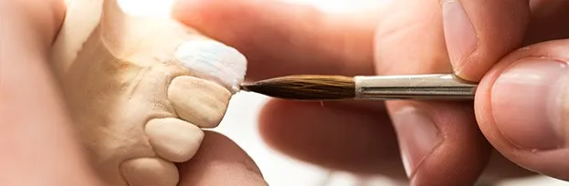 Ceramista aplicando porcelana con pincel sobre un modelo de resina dental durante la elaboración de una carilla feldespática